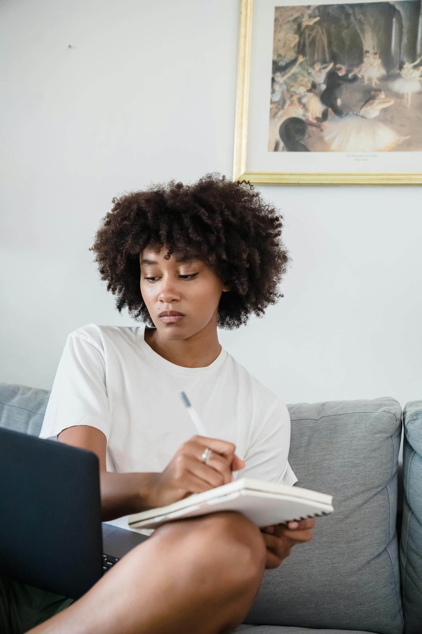 Young woman attentively studying at home using a laptop and notebook on a cozy sofa.