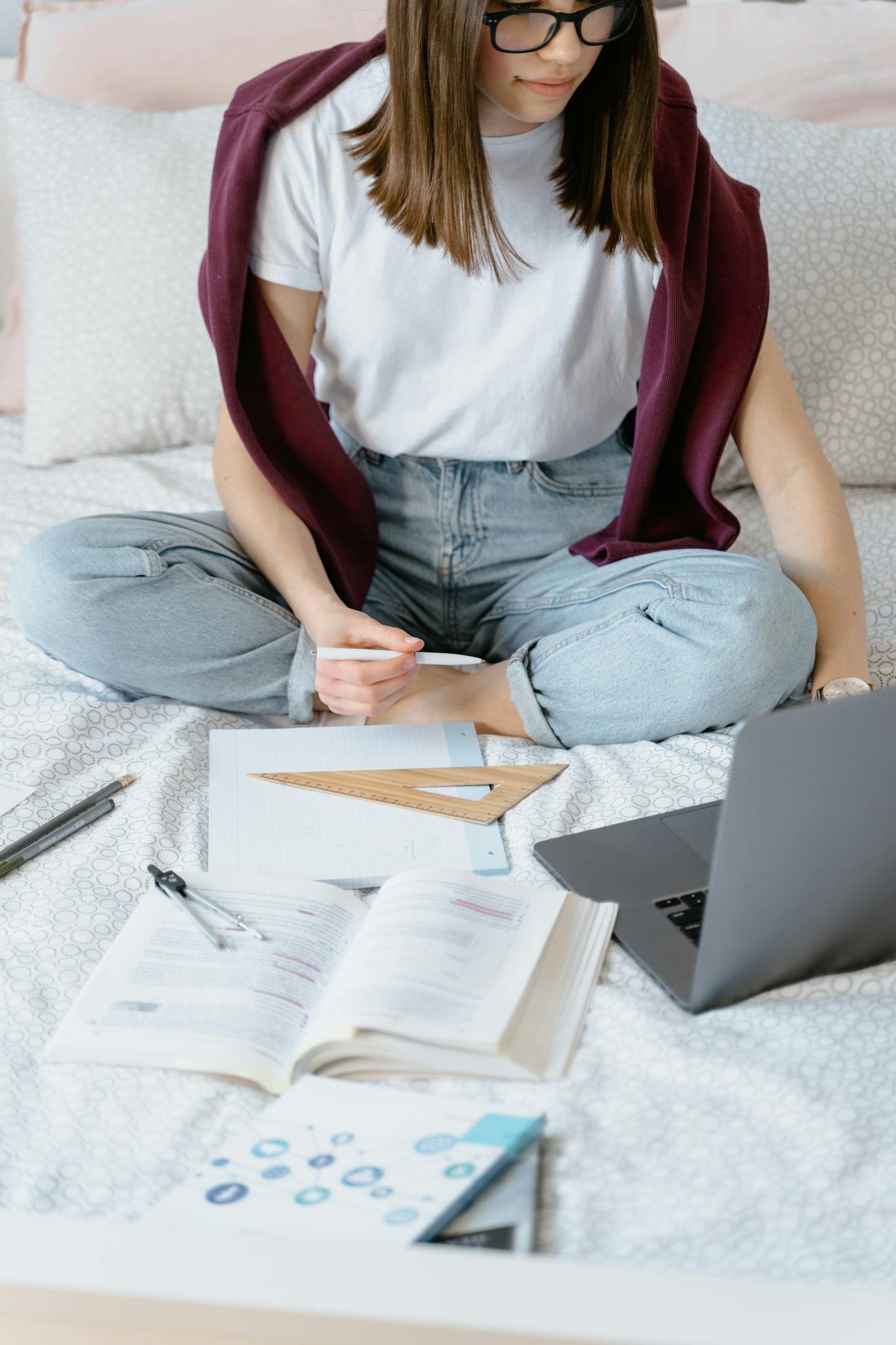 Young woman focused on studying from home with books and a laptop on her bed.